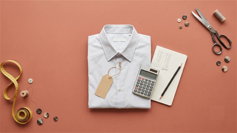 A folded white cotton shirt with a blank paper price tag, a calculator and a notebook of handwritten figures, with a measuring tape, scissors, buttons and thread on a terracotta background