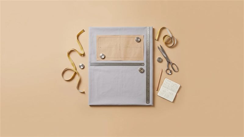 A grey cotton fabric sheet with a paper pattern rectangle on it and two steel rulers laid along its edges, with measuring tapes, scissors, a pencil and a small notebook on a warm sand background