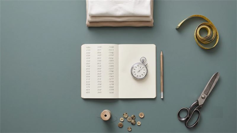 An open notebook of handwritten figures with a stopwatch on the page, a pencil, a stack of folded garments, a measuring tape, scissors, thread and buttons on a slate-green background