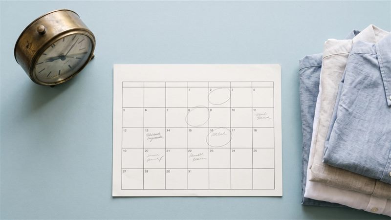 A paper wall calendar with a few days circled in pencil on a pale blue desk, with a brass desk clock to one side and a stack of folded shirts on the other